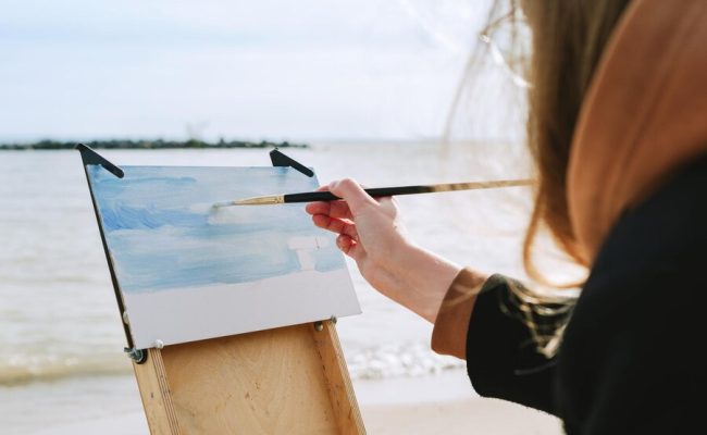 faceless-shot-of-a-female-drawing-on-a-beach-en-plein-air-artist_169478-1105